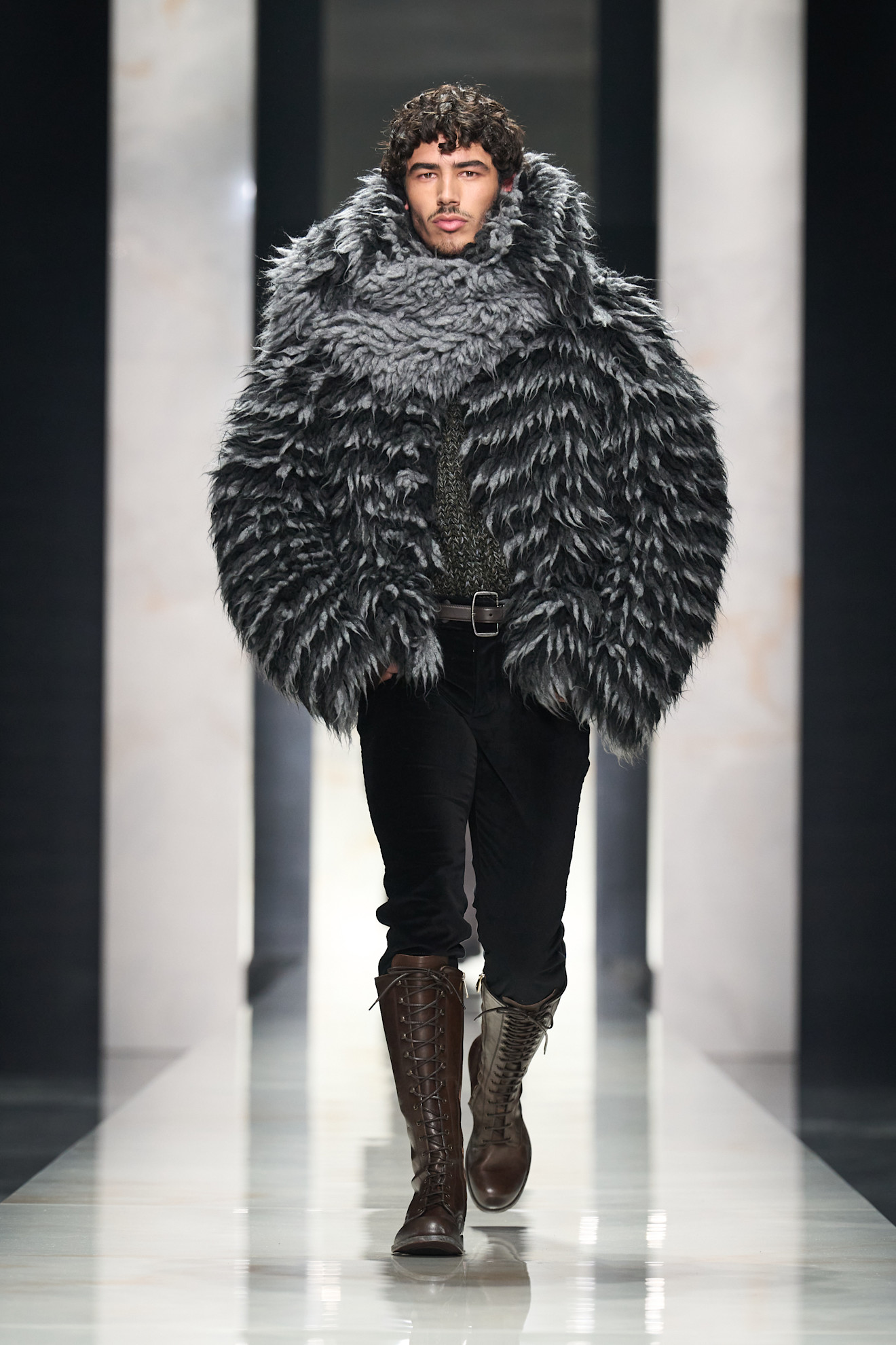 Мужская модель в объемном серо-черном меховом пальто с текстурой, сочетает с облегающими черными брюками и высокими коричневыми сапогами.