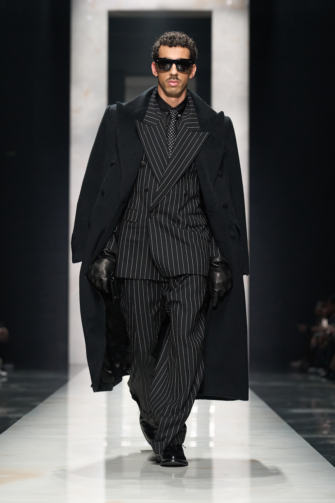 Элегантный образ с полосатым костюмом и длинным черным пальто, завершенный темными очками, излучает уверенность