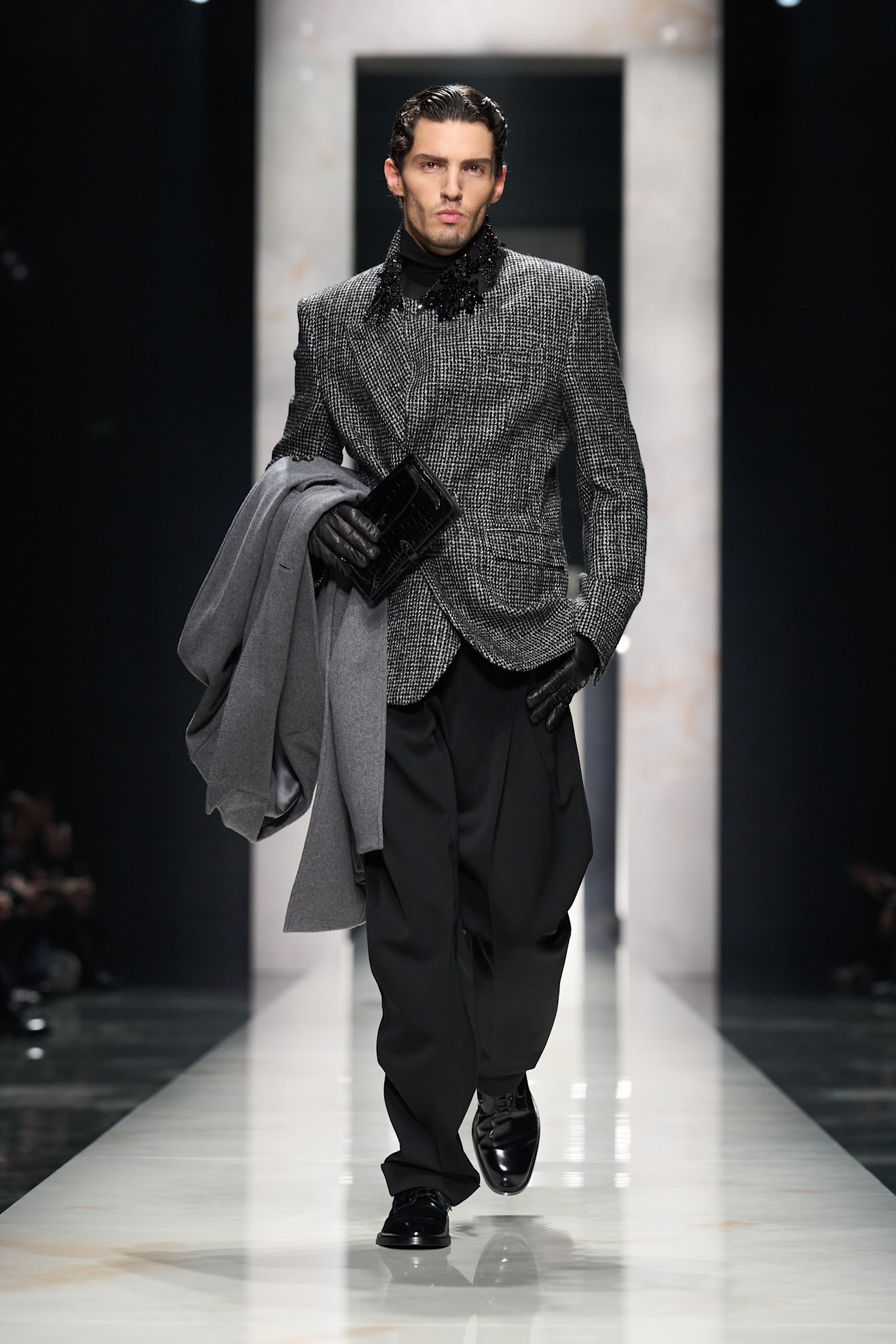 Строгий пиджак с блестящей текстурой и черной водолазкой, в руках модель держит серый плащ, создавая атмосферу утонченности