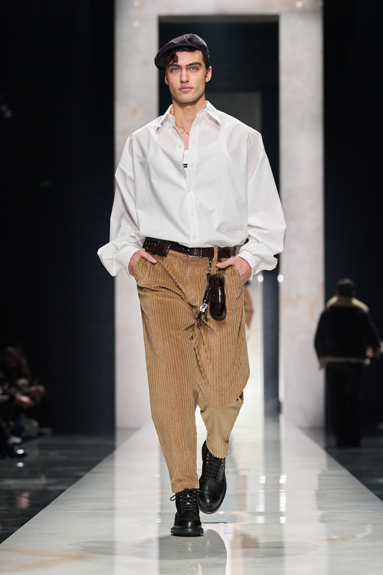 Объемная белая рубашка и коричневые вельветовые брюки создают стильный контраст, дополненный черной кепкой и ботинками