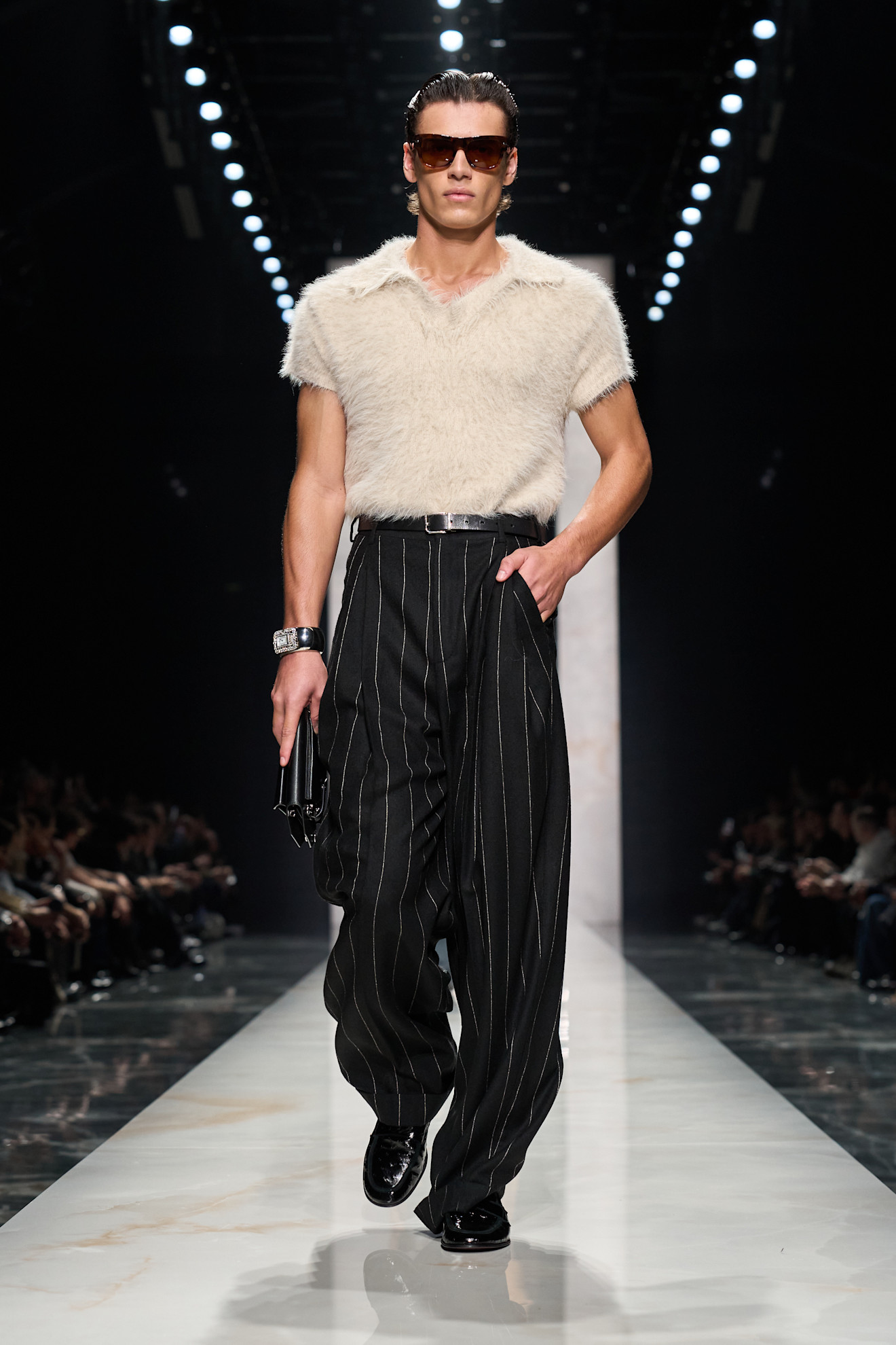 Модель в мягкой белой футболке и черных полосатых брюках, завершает образ стильными очками и лаконичным аксессуаром