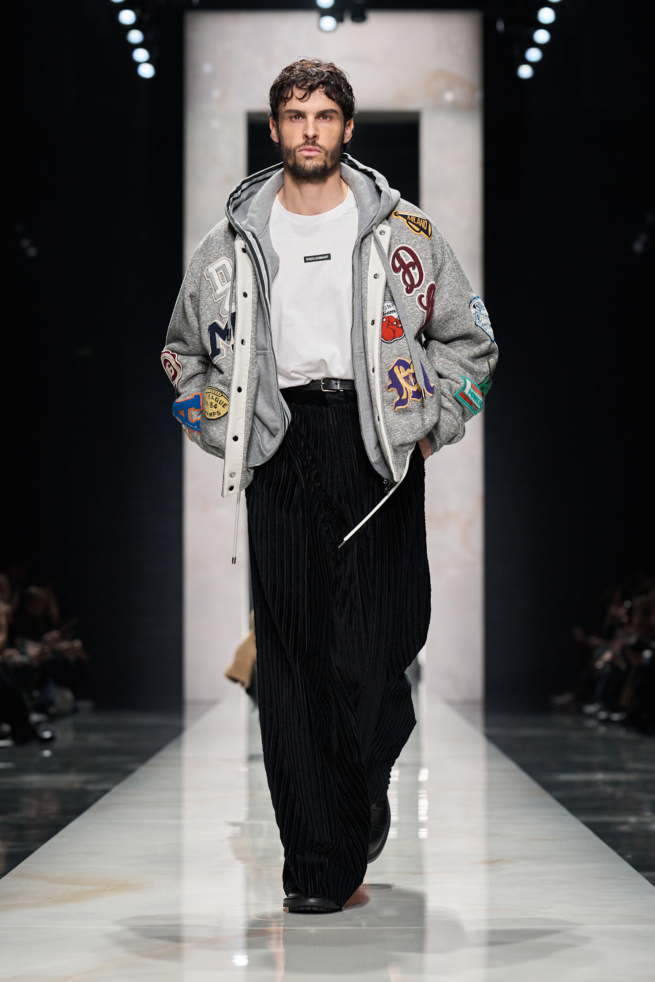 Объемный серый бомбер с нашивками, белая футболка и черные плиссированные брюки создают стильный уличный образ для осени 2026