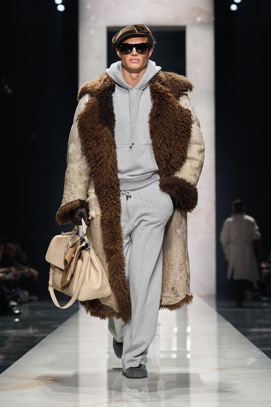 Объемный коричневый меховой плащ на сером спортивном костюме, дополненный стильной кепкой и сумкой в руках