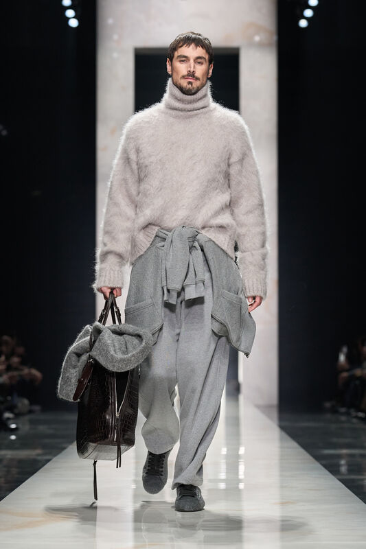 Пуховый светло-серый свитер с высоким воротом и серые брюки, в руках — стильная сумка и дополнительный свитер