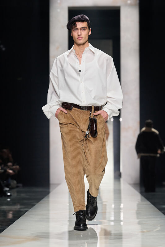 Объемная белая рубашка и коричневые вельветовые брюки создают стильный контраст, дополненный черной кепкой и ботинками