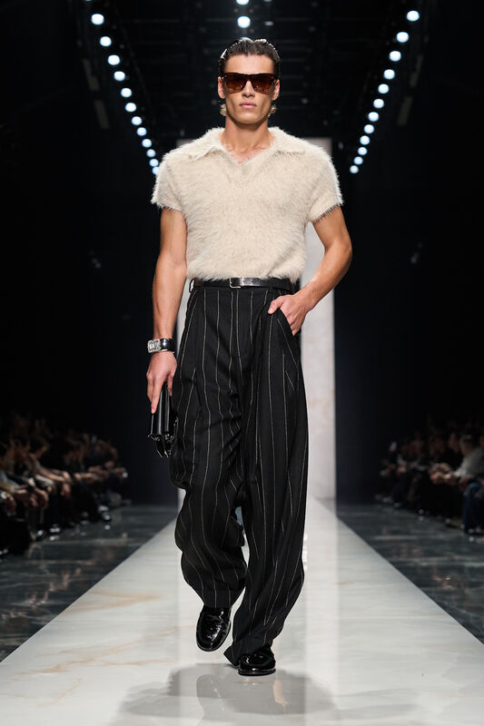 Модель в мягкой белой футболке и черных полосатых брюках, завершает образ стильными очками и лаконичным аксессуаром