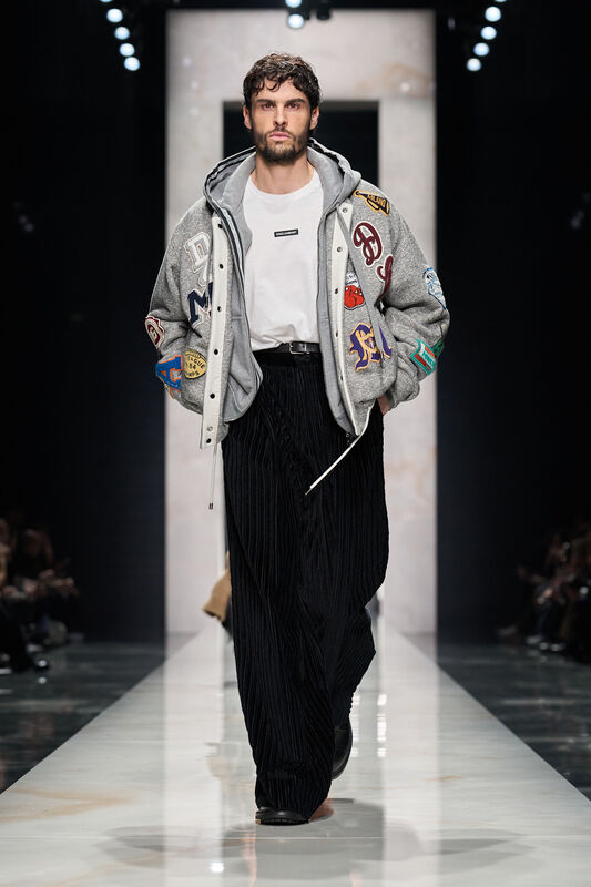 Объемный серый бомбер с нашивками, белая футболка и черные плиссированные брюки создают стильный уличный образ для осени 2026