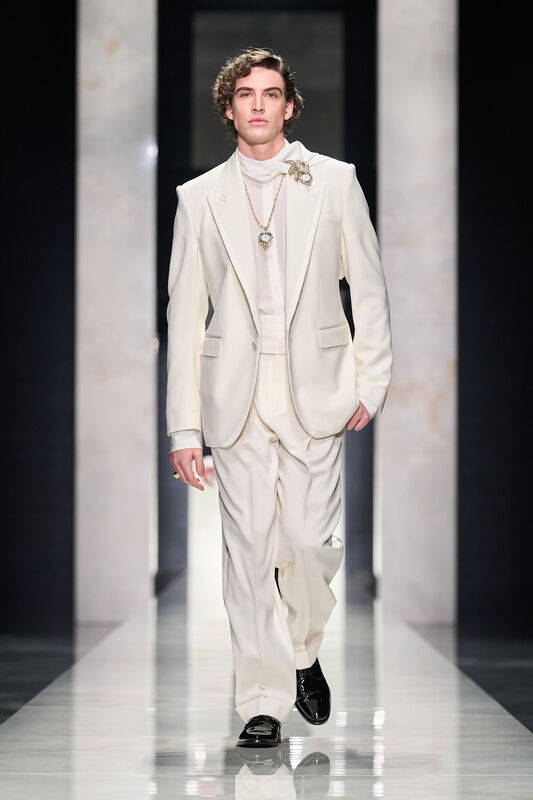 Светлый костюм с изысканными деталями, включая брошку и цепочку, создает атмосферу классической элегантности
