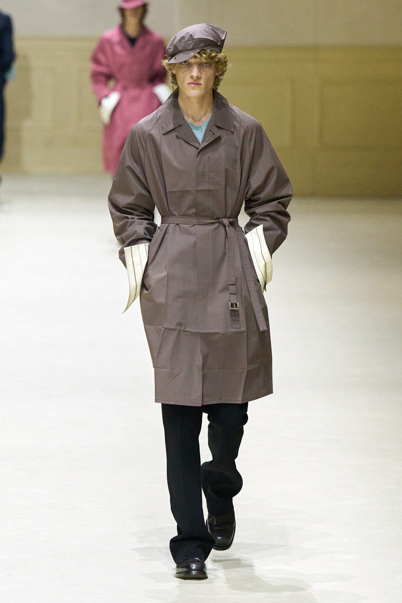 Модель в коричневом тренче с оригинальными рукавами, дополняет образ стильной кепкой и черными брюками