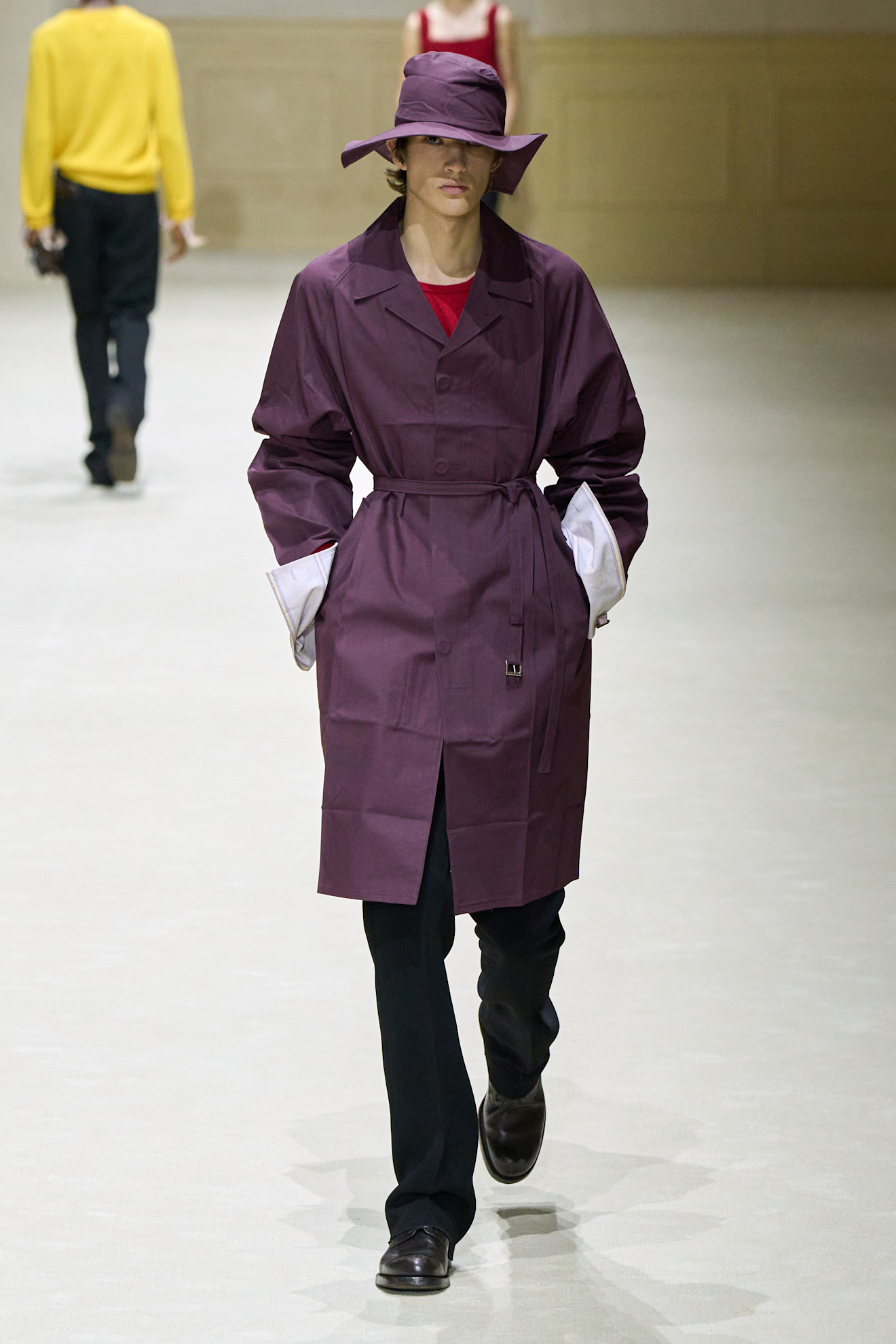 Модель в пурпурном тренче с шляпой, сочетая его с черными брюками и красным топом, привлекает внимание яркими цветами