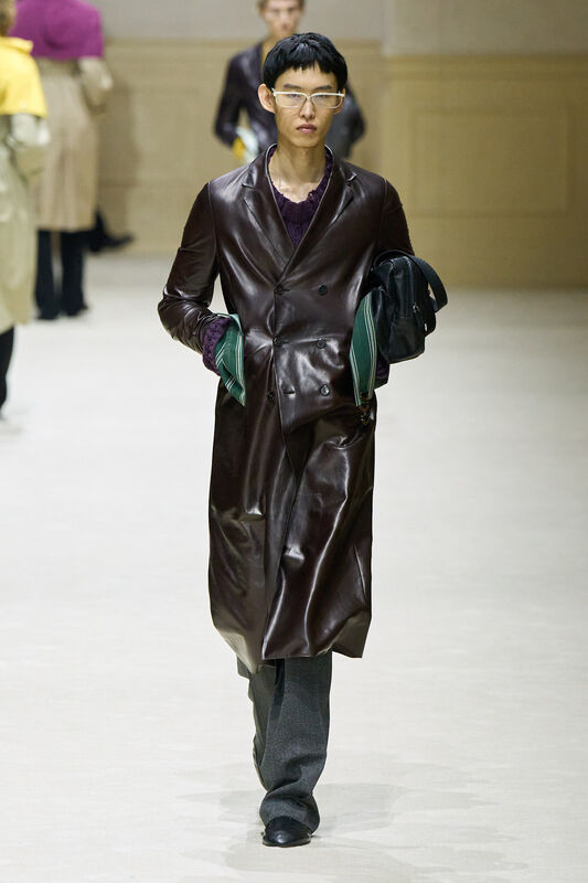 Модель в длинном коричневом пальто из лакированной кожи, подчеркивающем силуэт, с зеленым аксессуаром в руках