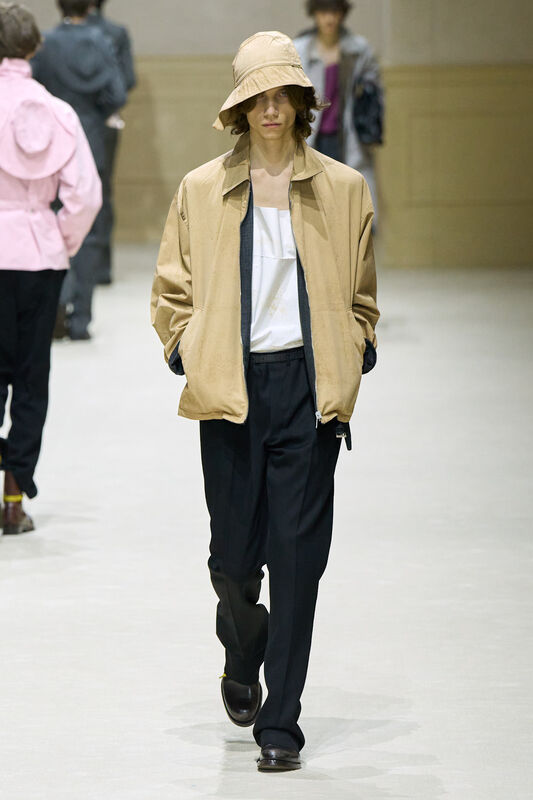 Модель в бежевом куртке и широких черных брюках, завершает образ стильной шляпой, создавая расслабленный вид