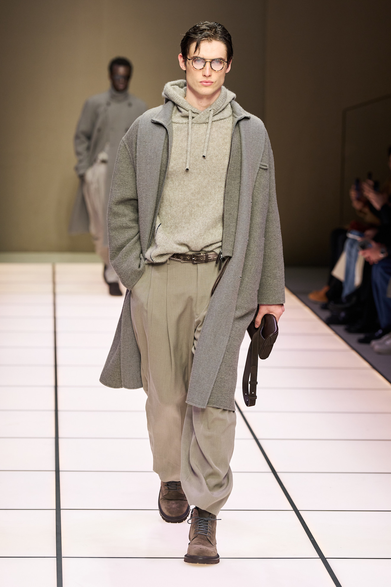 Молодой человек в сером пальто и свитере с капюшоном, дополненный стильными брюками и ботинками, излучает уверенность