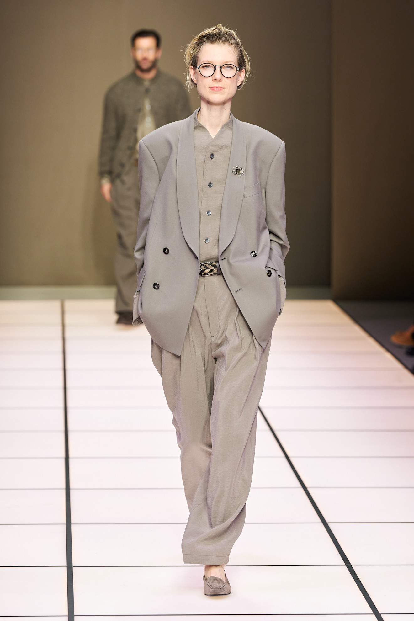 Модель в объемном сером костюме с широкими брюками и стильными очками, уверенно шагает по подиуму