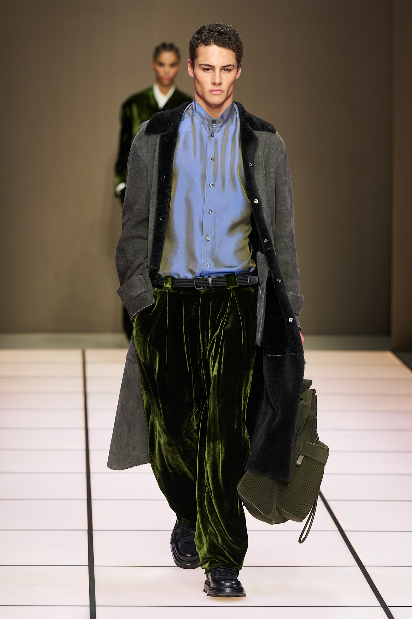 Модель в длинном сером пальто с меховой отделкой, сочетает его с яркой рубашкой и бархатными брюками глубокого зеленого цвета