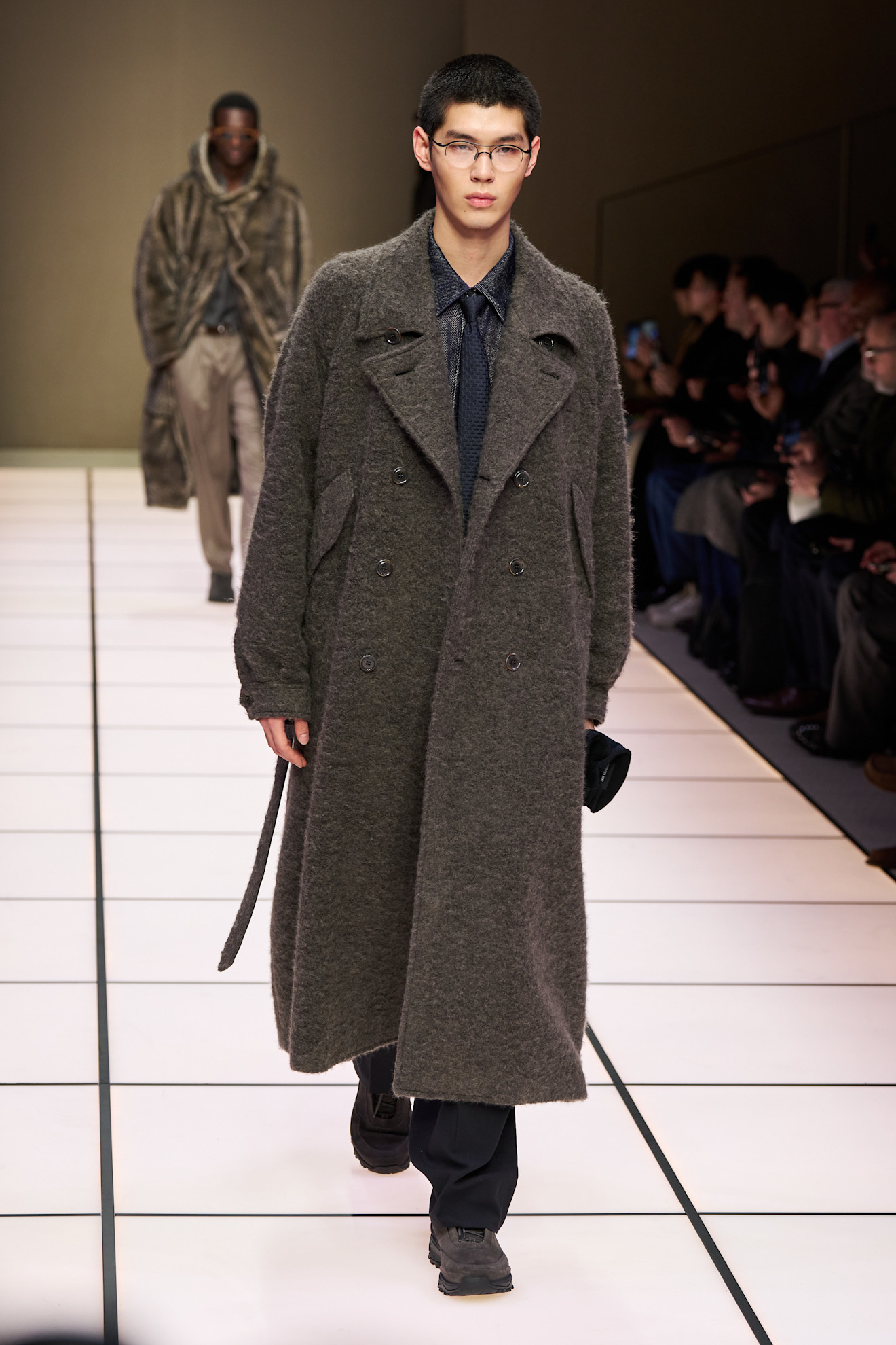 Модель в объемном сером пальто из мягкой ткани, дополненном строгой рубашкой и галстуком, уверенно шагает по подиуму