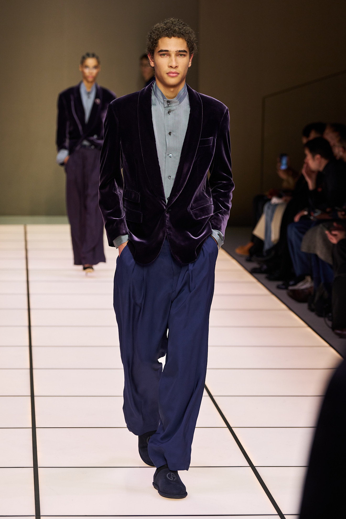 Молодой человек в бархатном пиджаке и светлой рубашке, сочетая элегантность с комфортом, уверенно идет по подиуму