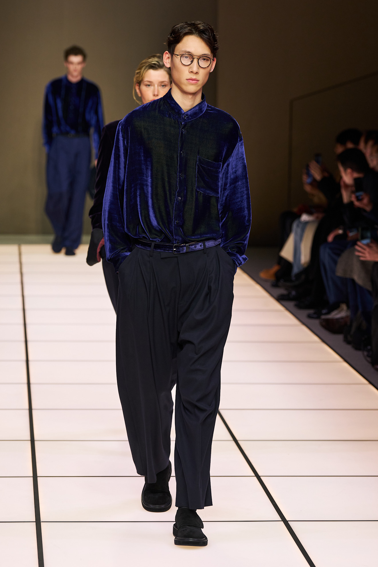 Модель в бархатной синей рубашке и широких брюках, уверенно шагает по подиуму, создавая атмосферу элегантности