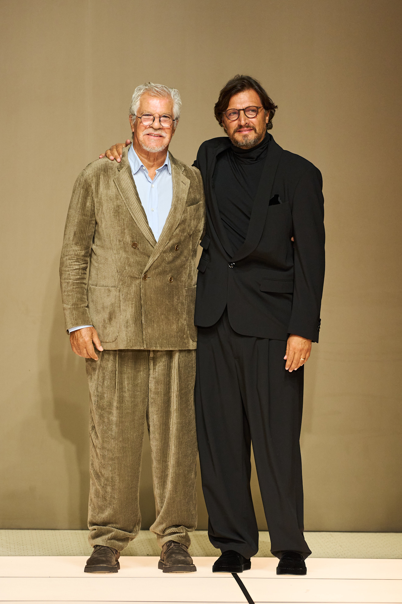 Два дизайнера на подиуме: один в коричневом вельветовом костюме, другой в черном смокинге, отражая стиль и креативность