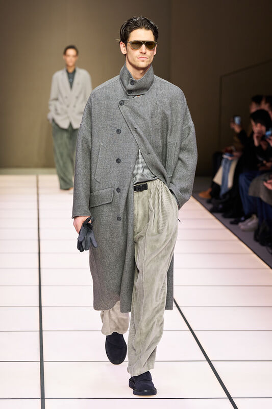 Модель в сером пальто с текстурой, подчеркивающим силуэт, сочетает его с полосатыми брюками и стильными очками