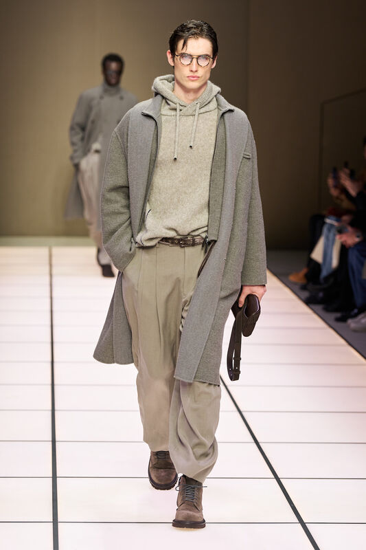 Молодой человек в сером пальто и свитере с капюшоном, дополненный стильными брюками и ботинками, излучает уверенность