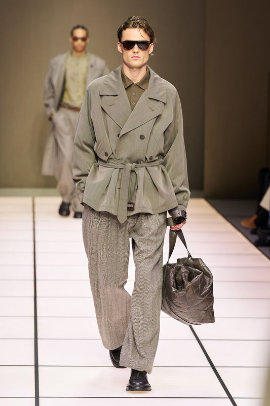 Модель в пиджаке с поясом и брюках в елочку, подчеркивающих силуэт, завершает образ с сумкой в тон