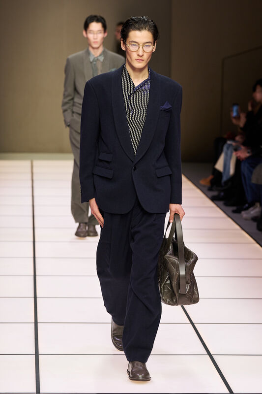 Молодой человек в темно-синем пиджаке и клетчатой рубашке, с сумкой в руках, создает образ современного делового стиля