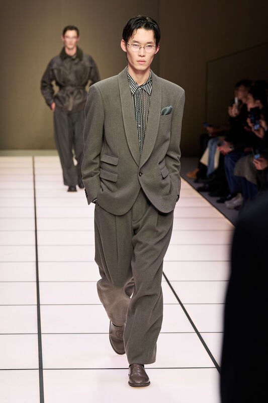 Модель в сером костюме с узорчатой рубашкой, уверенно шагает по подиуму, подчеркивая элегантность и стиль