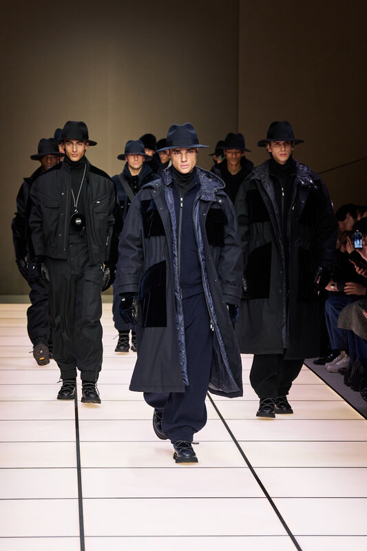 Группа моделей в черных и темно-синих нарядах с широкими шляпами, создающих атмосферу загадочности и стиля на подиуме