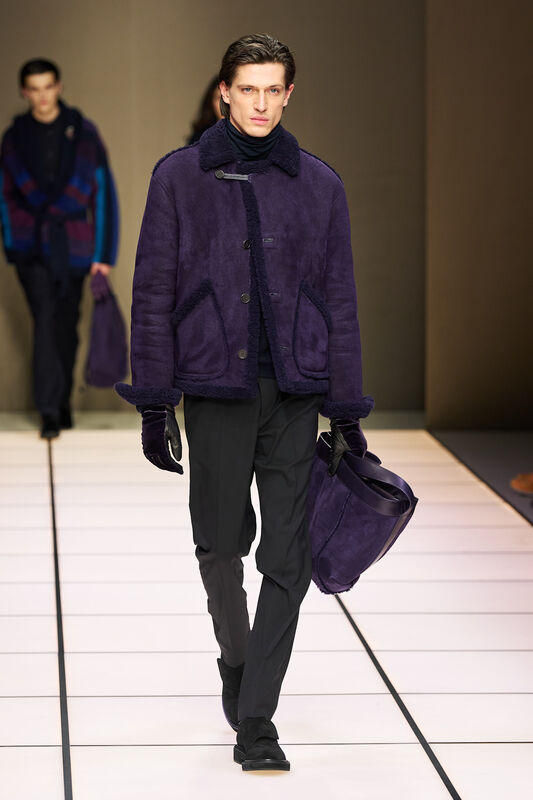Модель в насыщенном фиолетовом пальто с текстурой, дополненном черными перчатками и сумкой, уверенно шагает по подиуму