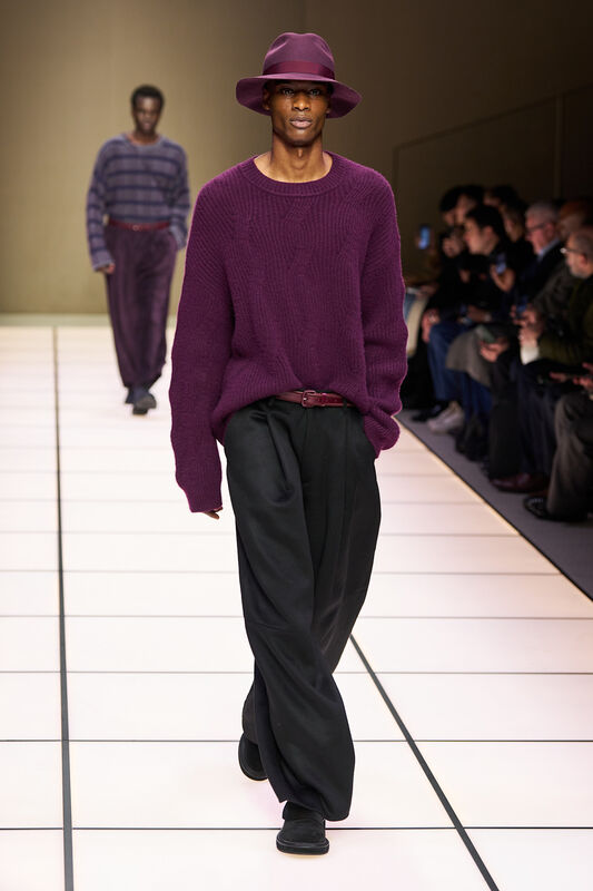 Модель в бордовом свитере и широких черных брюках с поясом, уверенно идет по подиуму, привлекая внимание