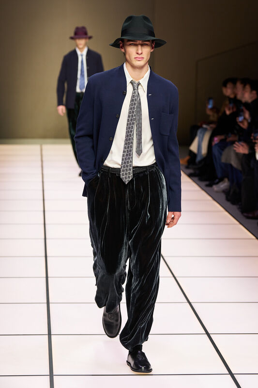 Модель в темно-синем пиджаке и бархатных брюках, завершает образ черной шляпой, создавая атмосферу классического стиля