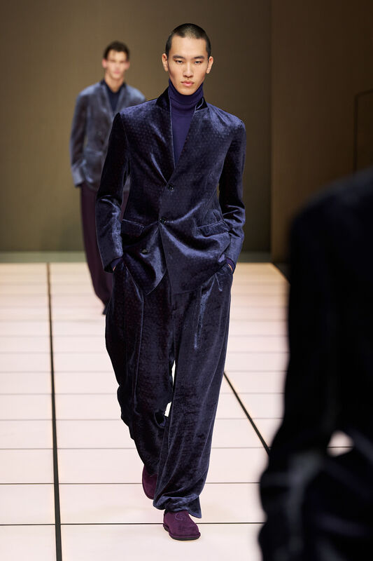 Модель в бархатном темно-синем костюме с широкими брюками и облегающим свитером, уверенно шагает по подиуму