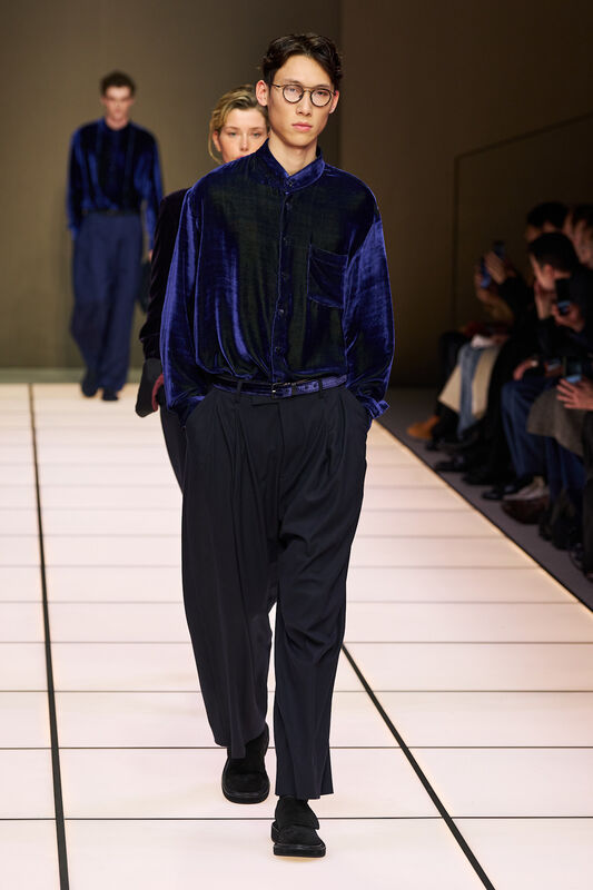 Модель в бархатной синей рубашке и широких брюках, уверенно шагает по подиуму, создавая атмосферу элегантности