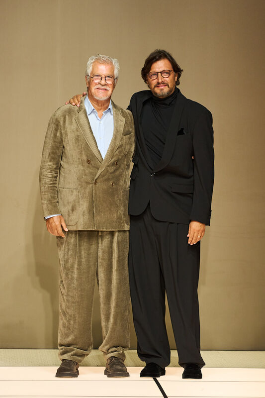 Два дизайнера на подиуме: один в коричневом вельветовом костюме, другой в черном смокинге, отражая стиль и креативность