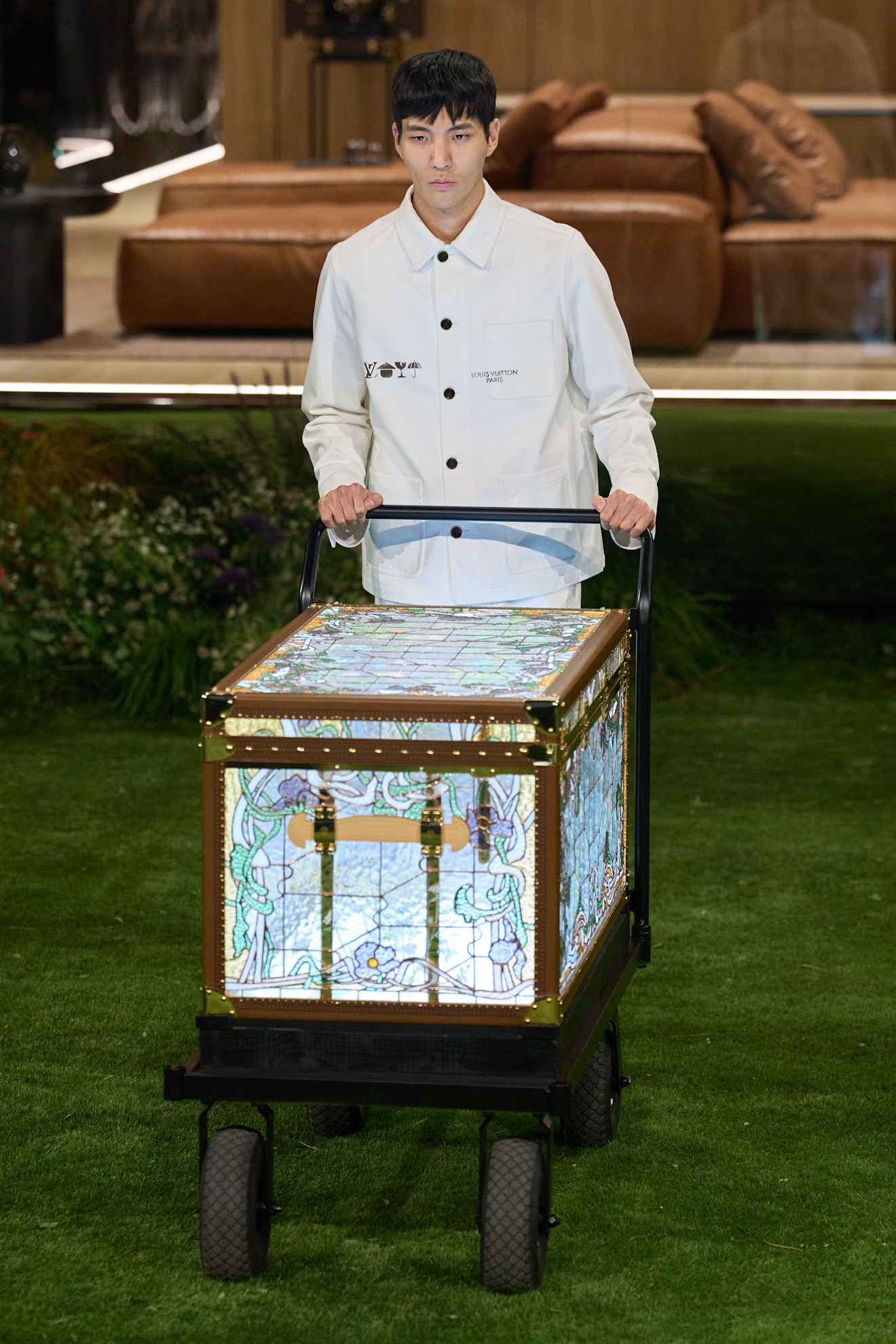 Модель в белой куртке с логотипом, толкает тележку с витражным чемоданом, символизируя креативный подход к путешествиям