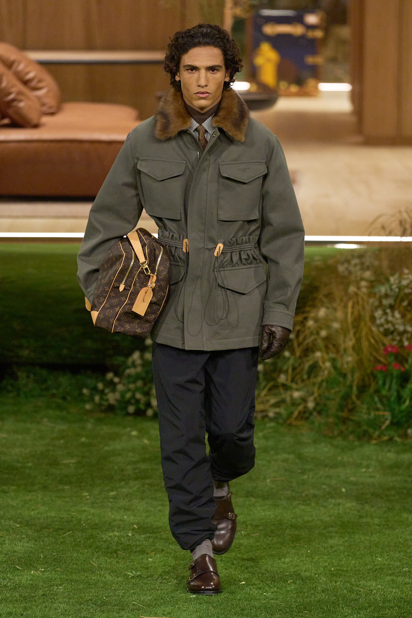Мужчина в стильном парке с меховым воротником и кожаных перчатках, держит сумку Louis Vuitton на фоне зелёного газона