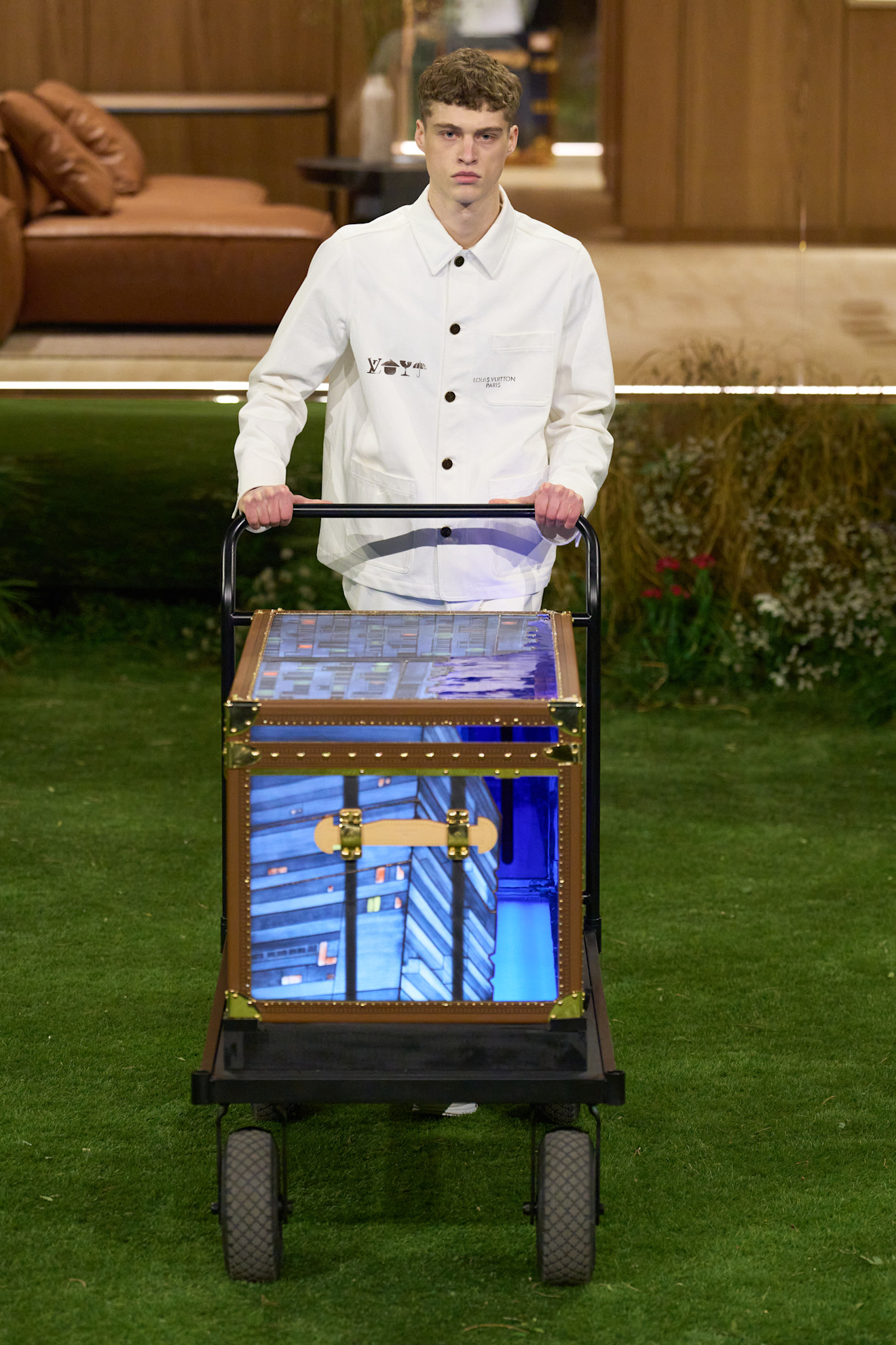 Модель в белом куртке с логотипами Louis Vuitton, толкает тележку с ярким чемоданом, на фоне зелёного газона
