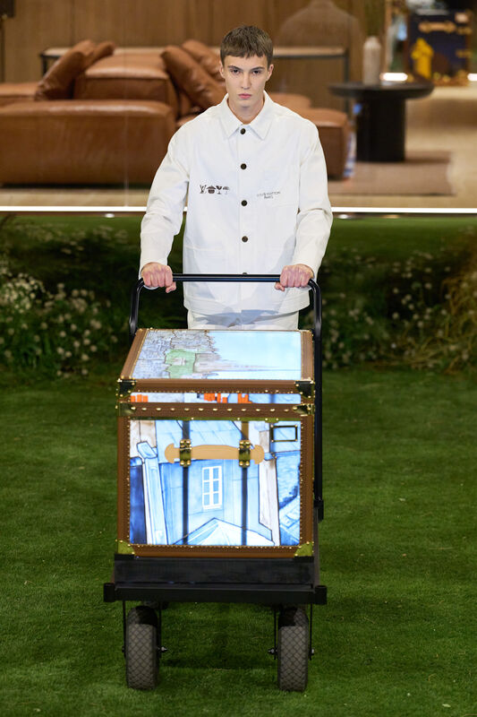 Модель в белом куртке с карманами, толкает тележку с ярким чемоданом, на фоне зелёного газона и уютной обстановки