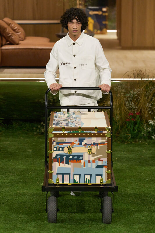 Модель в белом куртке с логотипом, толкает тележку с ярким чемоданом, отражая игривый дух коллекции