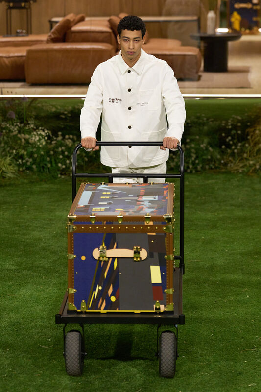 Молодой человек в белом комбинезоне управляет тележкой с ярким чемоданом, украшенным графическим принтом