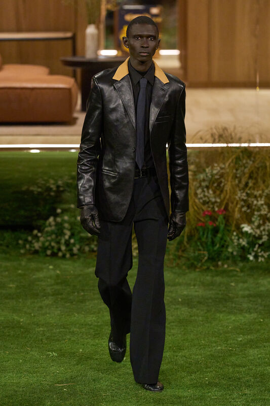 Модель в черном кожаном пиджаке с контрастным воротником, уверенно шагает по зелёному газону, создавая эффектный силуэт