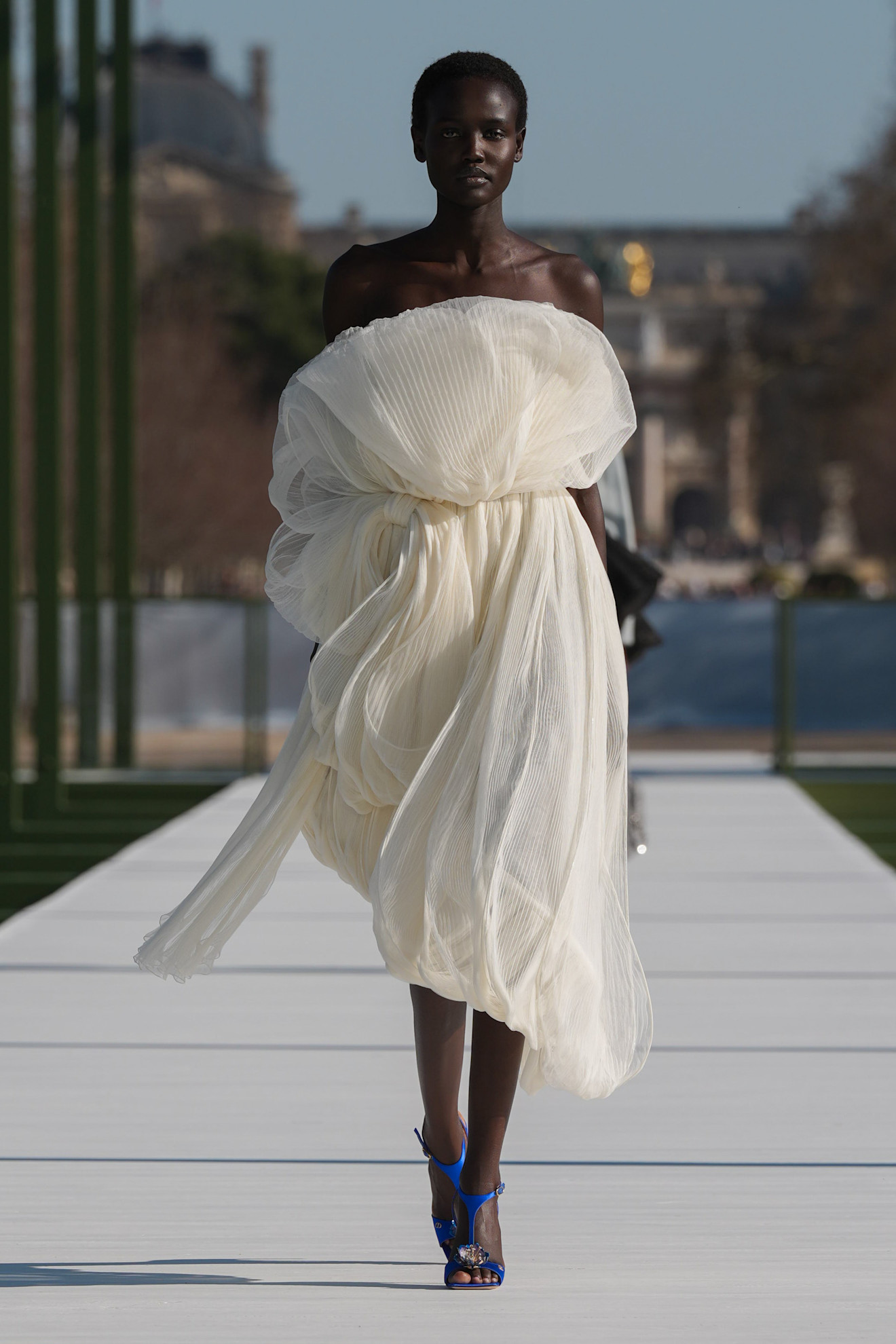 Легкое белое платье с объемными складками, подчеркивающее силуэт, дополнено яркими синими босоножками