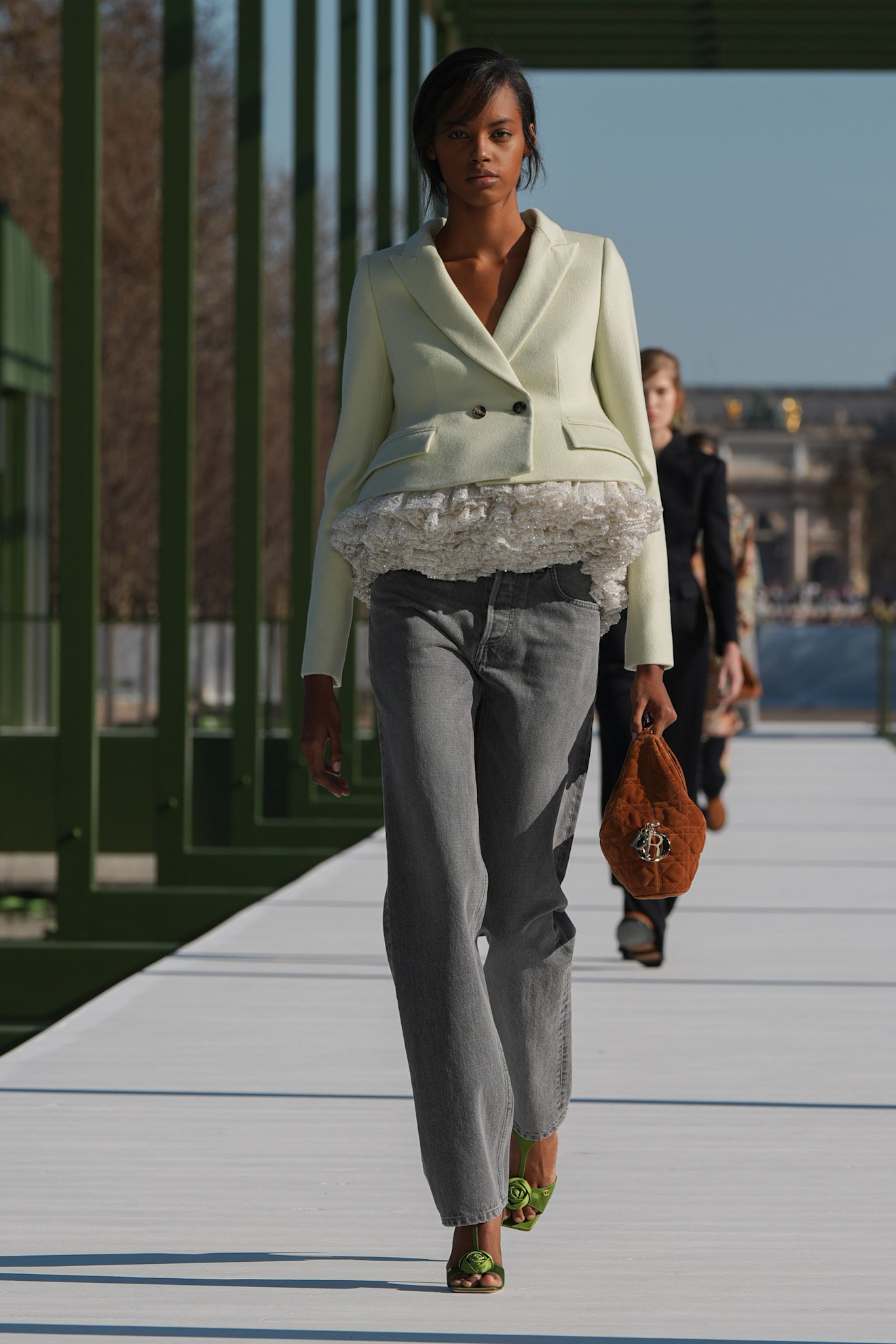 Пастельный жакет с рюшами и серые джинсы, яркие сандалии, уверенная походка на фоне зелёных конструкций