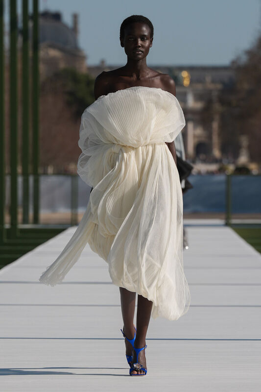 Легкое белое платье с объемными складками, подчеркивающее силуэт, дополнено яркими синими босоножками