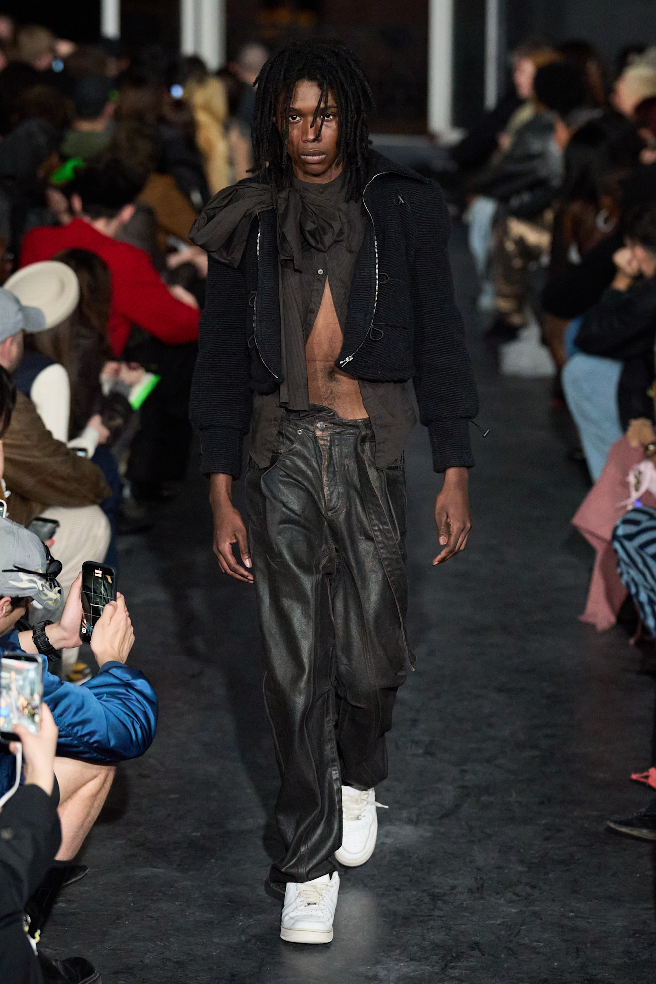 Модель в черном текстурированном жакете и широких брюках из кожи, уверенно шагает по подиуму, создавая атмосферу уличного стиля