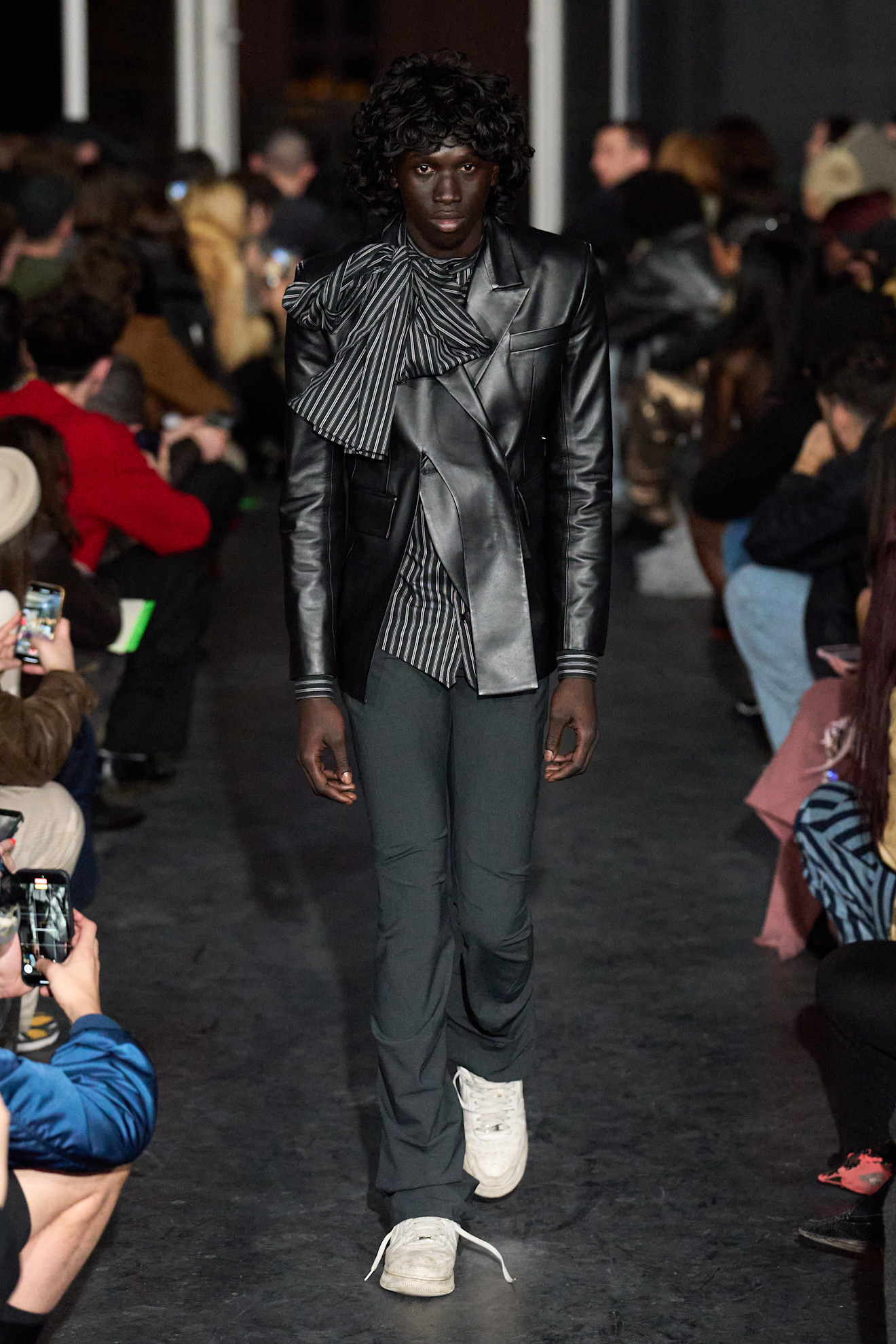 Модель в черном кожаном пиджаке с полосатым элементом, сочетает строгий стиль с игривыми деталями, уверенно шагая по подиуму