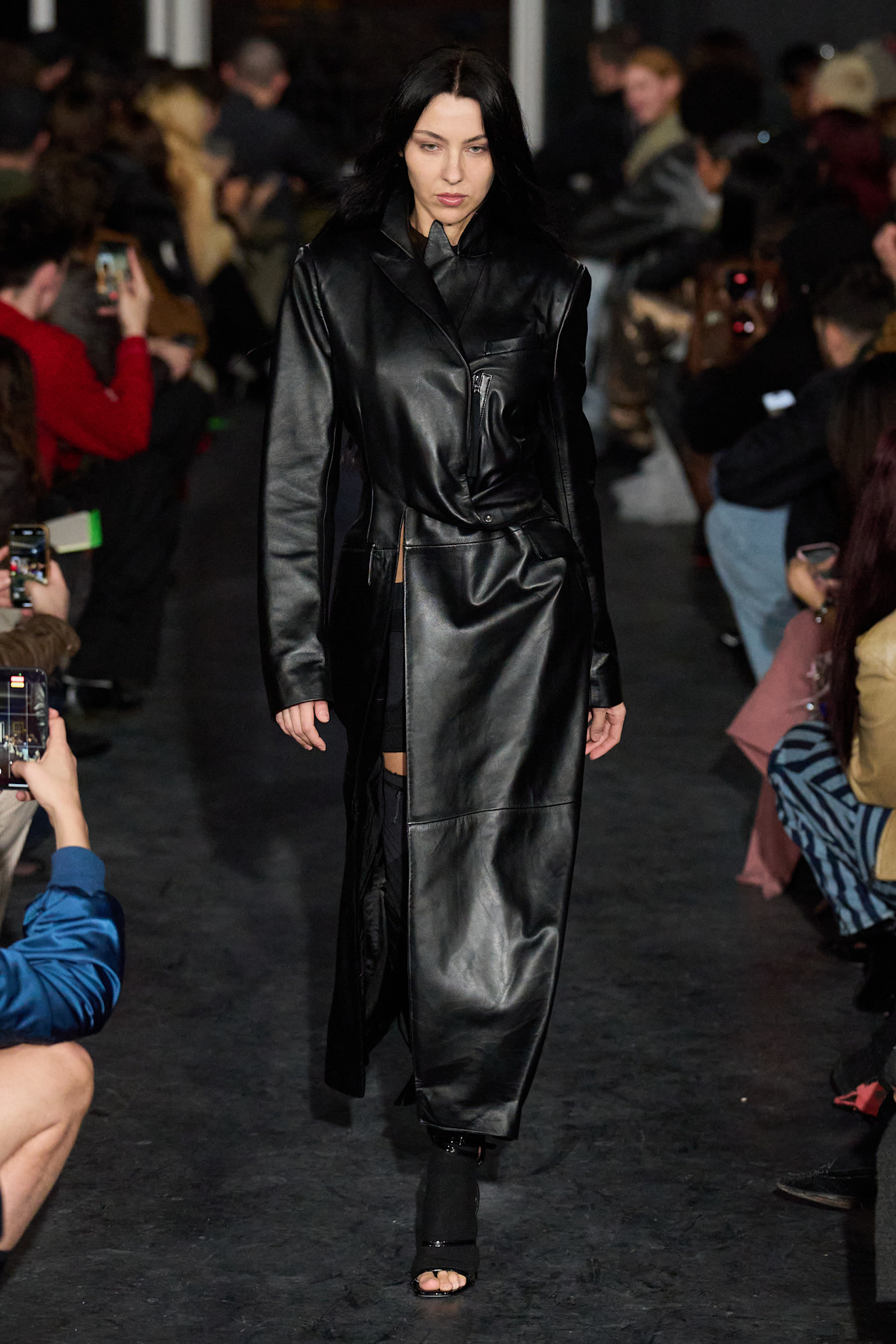 Модель в длинном кожаном пальто с асимметричным кроем, уверенно шагает по подиуму, создавая атмосферу загадочности