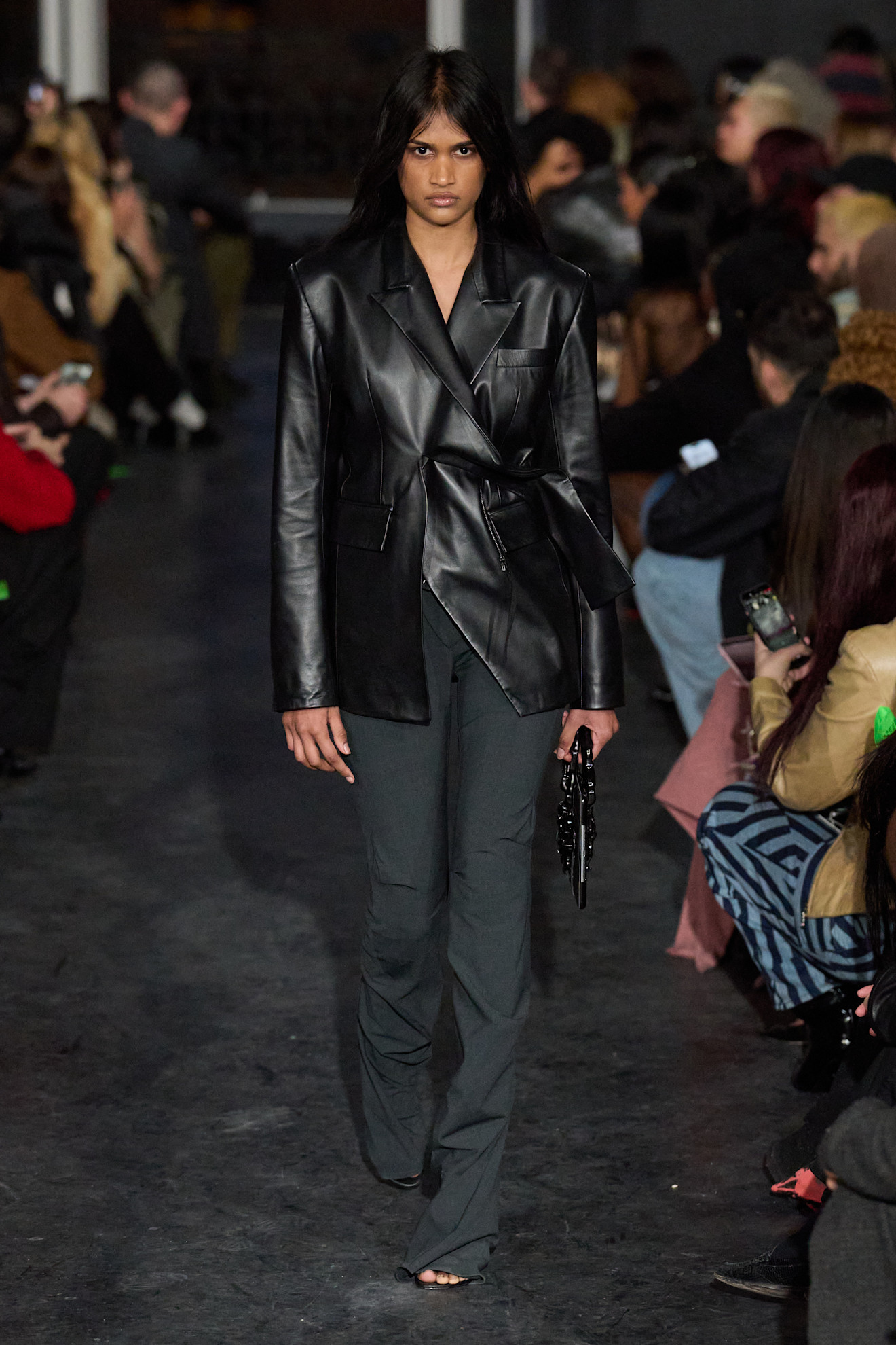 Кожаный пиджак с поясом и брюки в тон, создают стильный образ, подчеркивающий силуэт модели на фоне зрителей