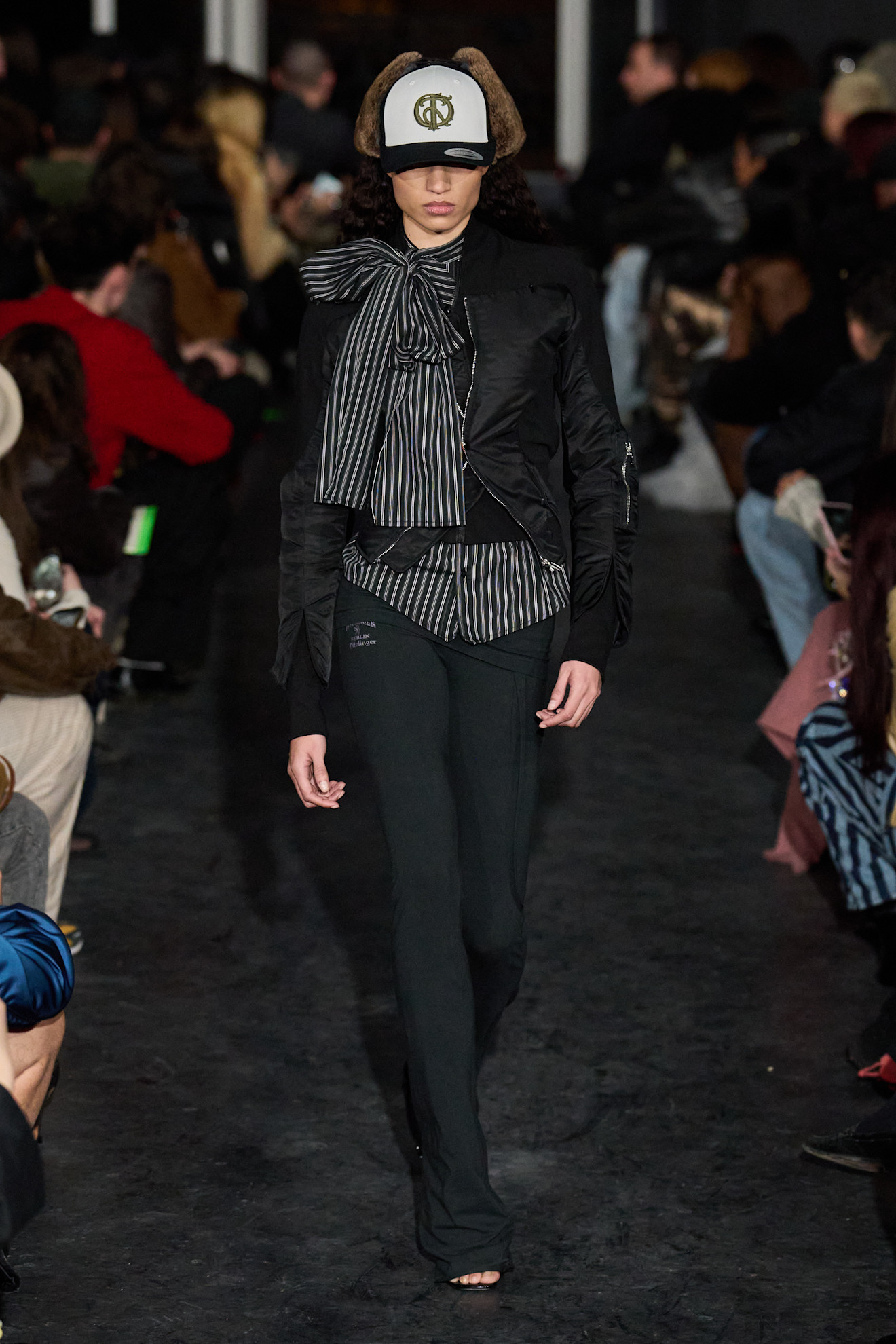 Модель в черной куртке с объемным бантом и полосатой блузкой, дополняет образ кепкой с меховыми ушами